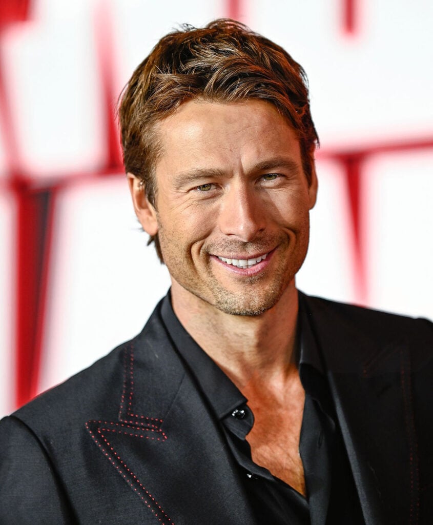 The Running Man London premiere red carpet, Glen Powell, Brioni custom suit, red silk-trimmed lapel, classic menswear tailoring, all-black suit red carpet