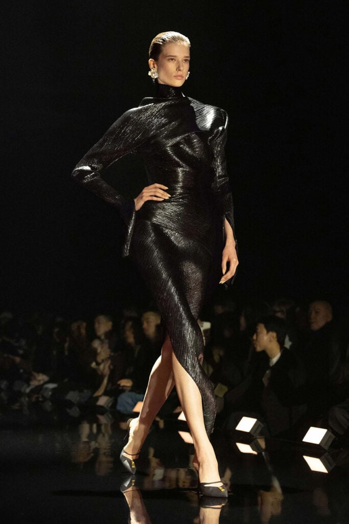 Model walks the runway in a black spiral-cut Schiaparelli Fall 2026 pleated satin dress with dramatic sleeves.