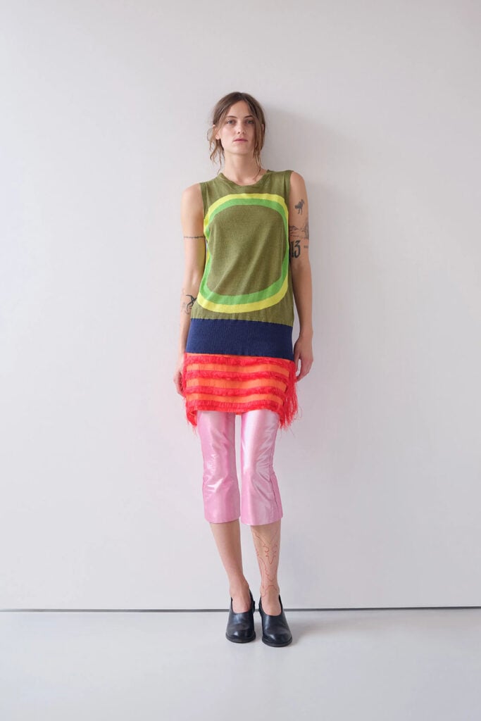Edward Cuming Spring 2026 runway look featuring the olive knit top, fringed skirt and glossy pink capri trousers.