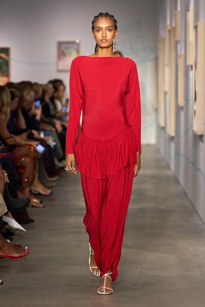 Ashlyn Spring 2026 red jumpsuit featuring a draped peplum and relaxed trousers on the runway.