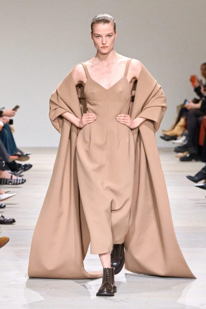 Emilia Wickstead runway look featuring a fitted taupe dress and cape styling.