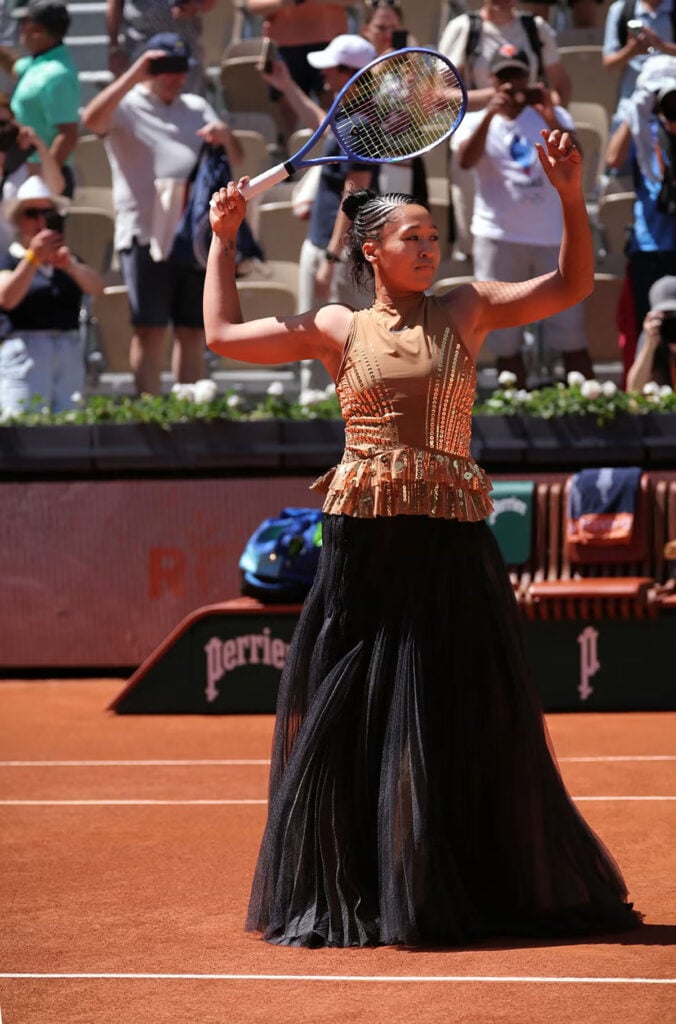 Naomi Osaka walking onto court in a bronze Nike x Germanier tennis outfit with flowing train at Roland-Garros 2026