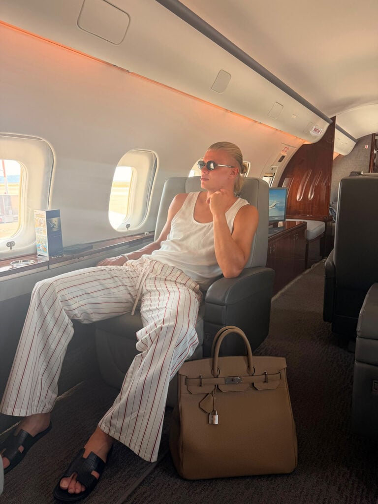 Erling Haaland wearing Dolce & Gabbana Spring 2026 striped trousers on a private jet with a Hermès HAC 40 bag and Hermès Izmir sandals.