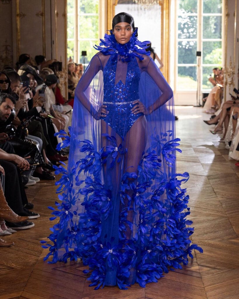 A model presents a cobalt blue Georges Chakra Fall 2026 Couture look with a feather-trimmed tulle cape on the runway.