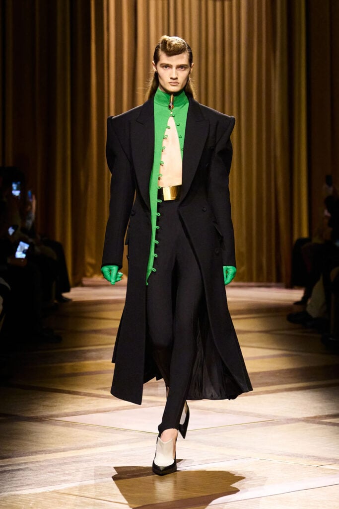 Mugler Fall 2026 runway look featuring a black extended-shoulder coat, green high-neck top and tailored trousers.