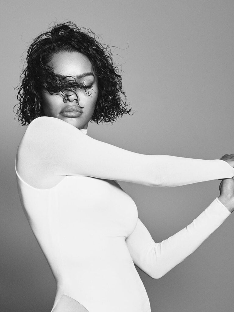 Teyana Taylor models a white second-skin Alaïa gown with curved hip cut-outs in a black-and-white campaign portrait.