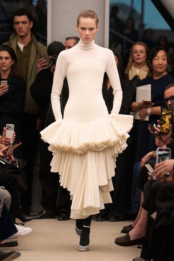 Alaïa Fall 2026 ivory high-neck dress with a fitted bodice and layered asymmetrical pleated skirt on the runway.