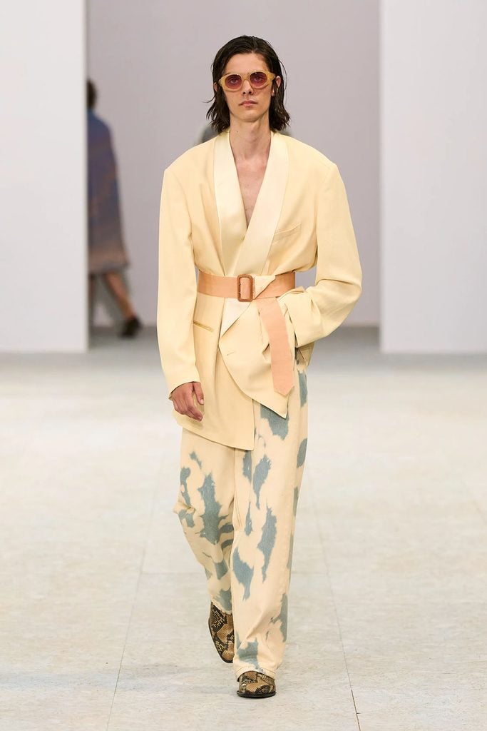 Dries Van Noten Spring 2027 menswear runway look featuring a cream shawl-collar jacket, peach belt and bleached jeans.