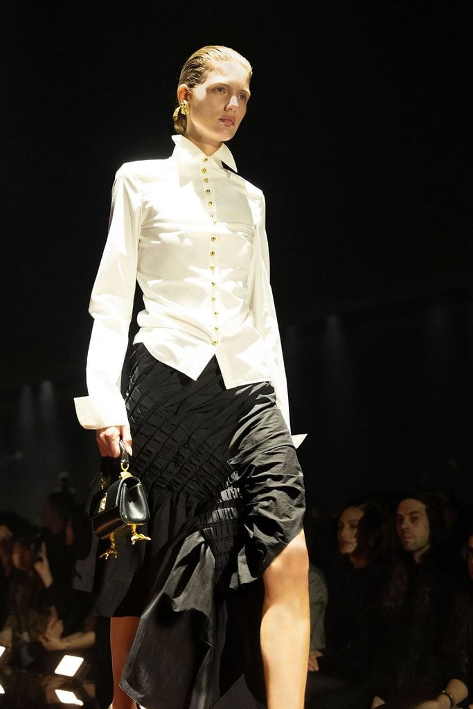 Schiaparelli Fall 2026 runway look featuring a fitted white buttoned shirt and asymmetric black pleated taffeta skirt.