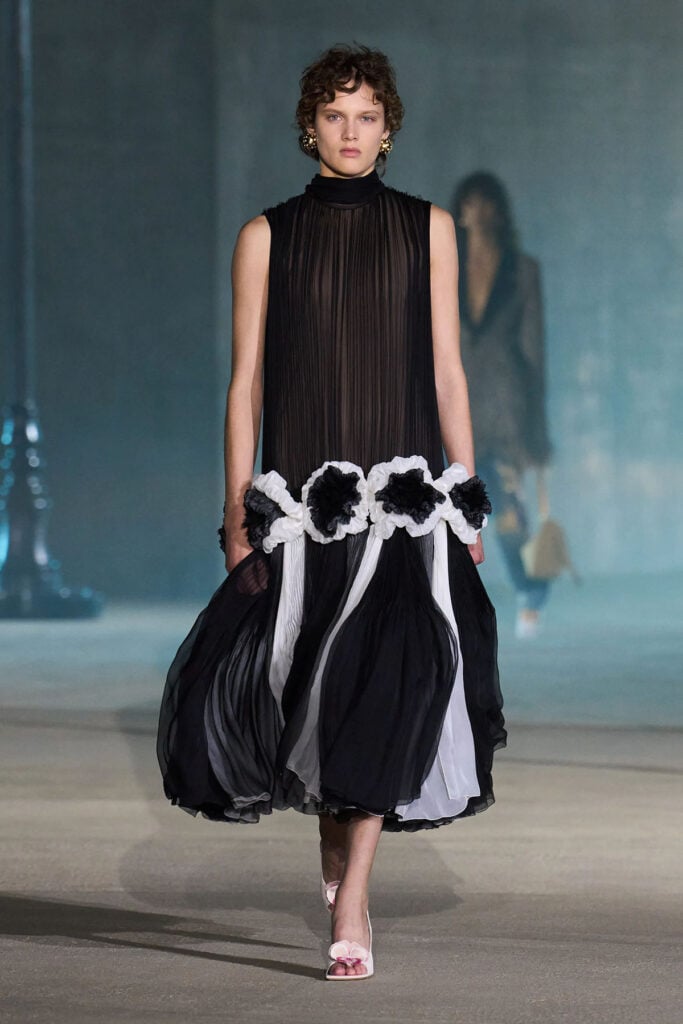 A model wears the black pleated Dior Resort 2027 dress with white tulle flowers on the runway.