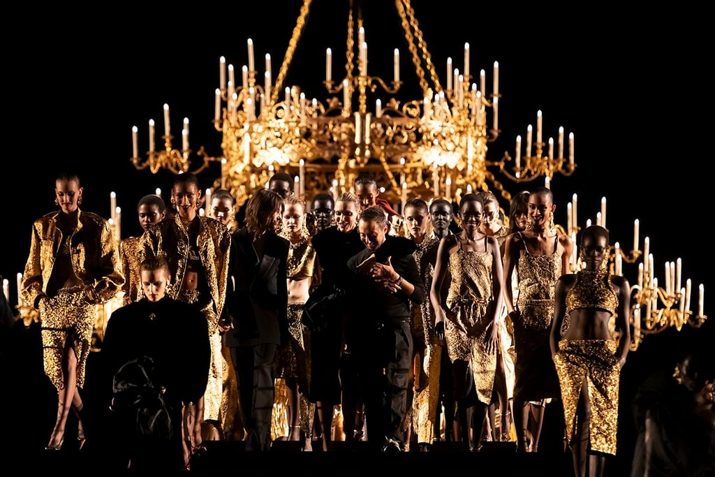 Anthony Vaccarello dressed in black surrounded by models in gold looks during the Saint Laurent Spring 2027 finale in Paris.