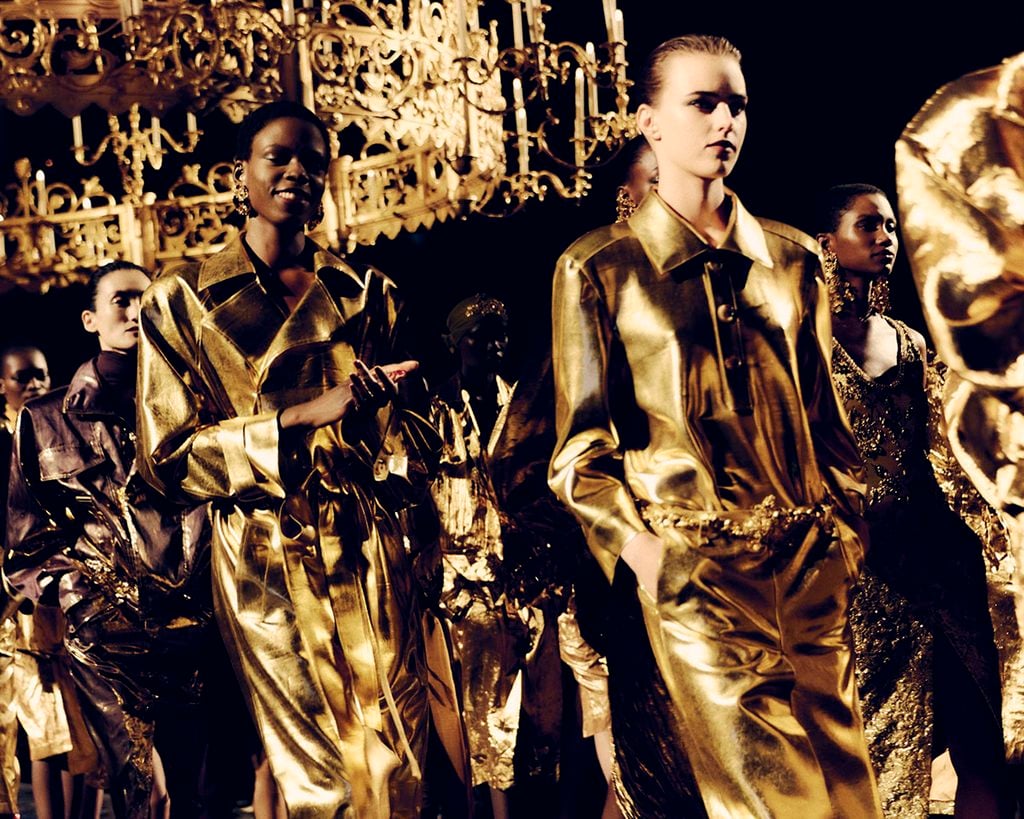 Models wearing metallic gold Saint Laurent looks beneath chandeliers during the Spring 2027 runway show in Paris.