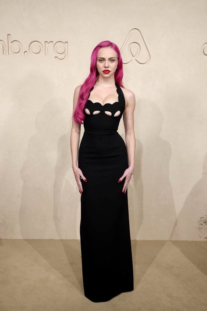 Adéla poses in a floor-length black gown with cut-outs beneath the bust, bright pink hair and red lipstick at the 2026 Airbnb.org Gala in Los Angeles