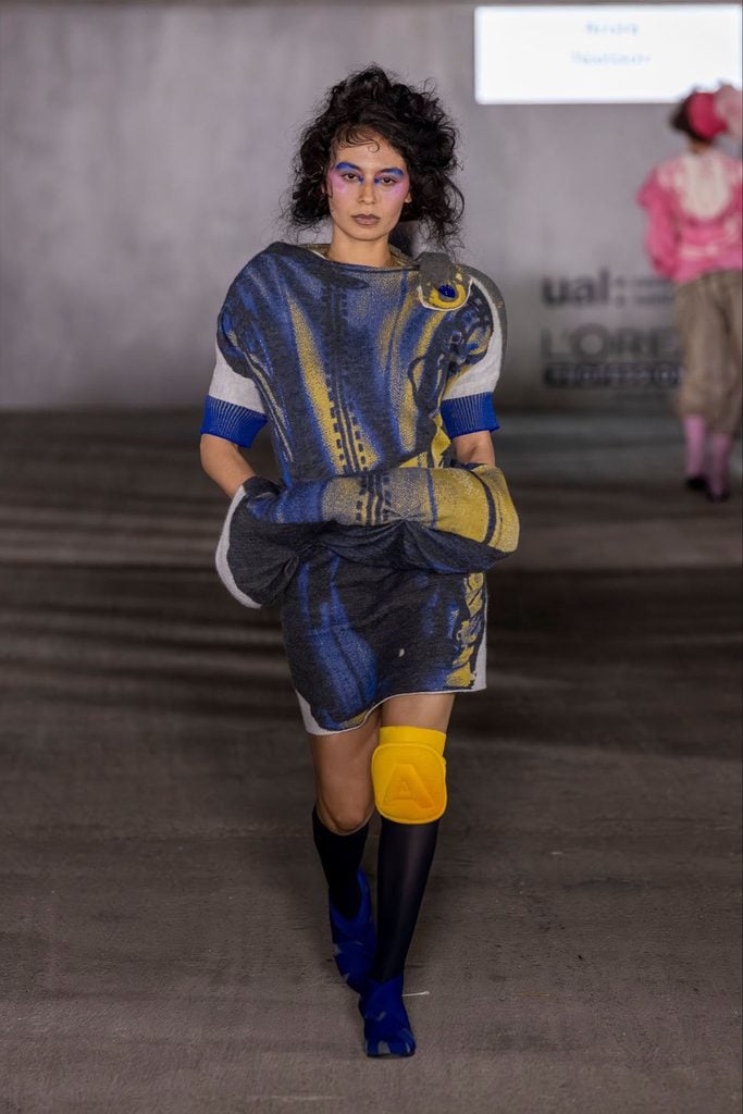Arora Nielson Fall 2026 Central Saint Martins runway look featuring sculptural blue-and-yellow knitwear, exaggerated padded hips and a yellow knee pad.