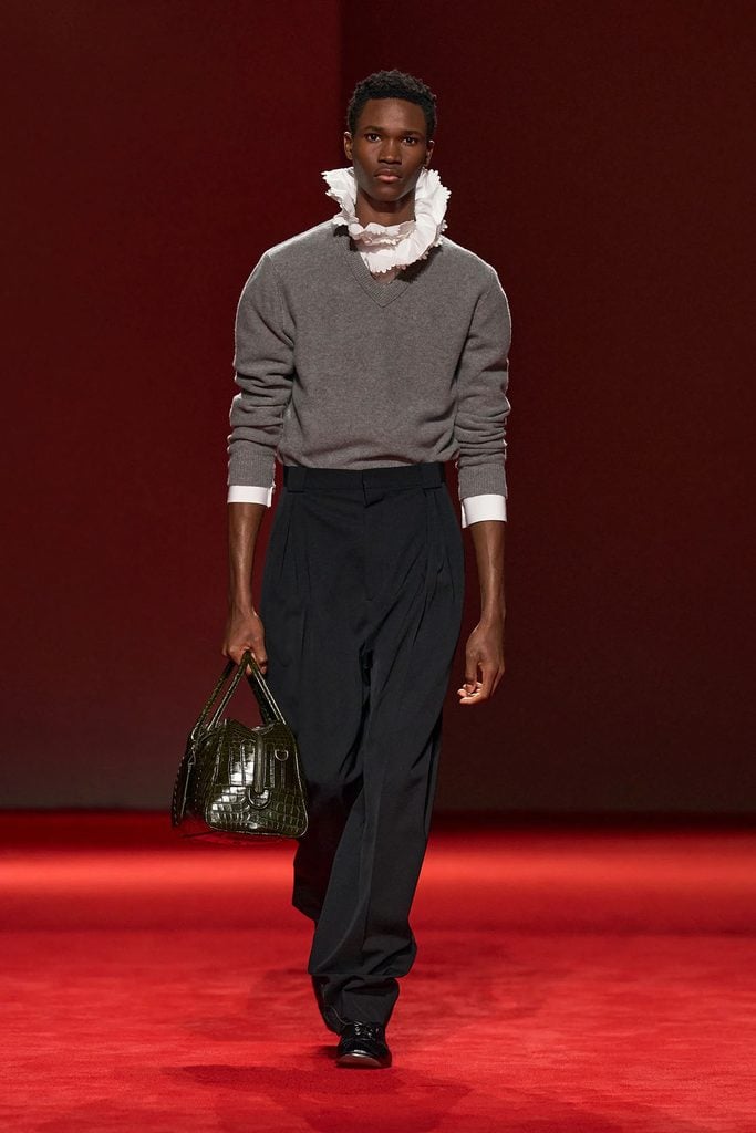 Bottega Veneta Fall 2026 runway look featuring a grey V-neck jumper, dramatic white ruffled collar, black tailored trousers and a dark green handbag.
