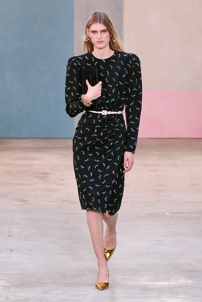 Model wearing a black shoe-print dress with a white belt and gold pumps on the Carolina Herrera Fall 2026 runway.