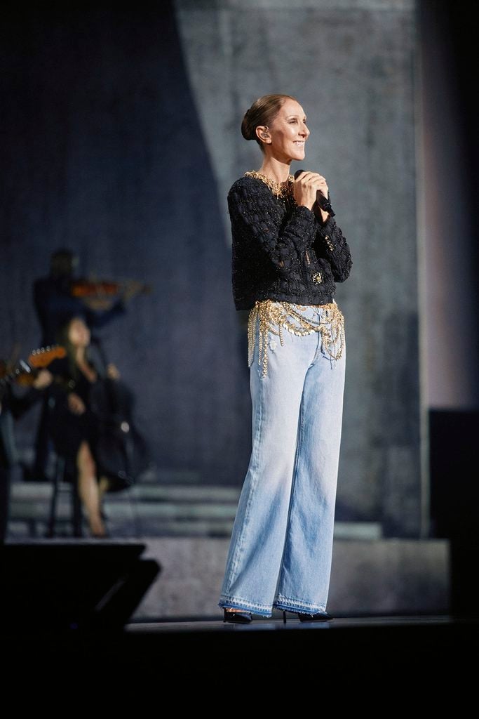 Céline Dion smiles onstage in Paris wearing a Chanel Fall 2026 Haute Couture jacket with cascading gold chain details over relaxed denim jeans.