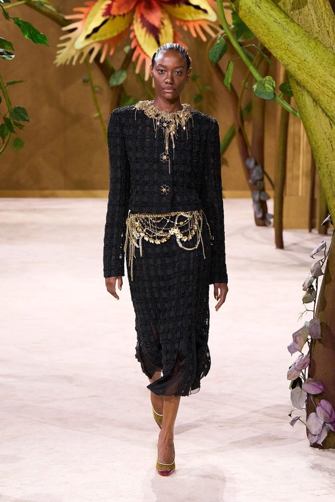 Chanel Fall 2026 Haute Couture runway model wearing a black tweed jacket and matching skirt with intricate gold chain braiding and embellishments in Paris.