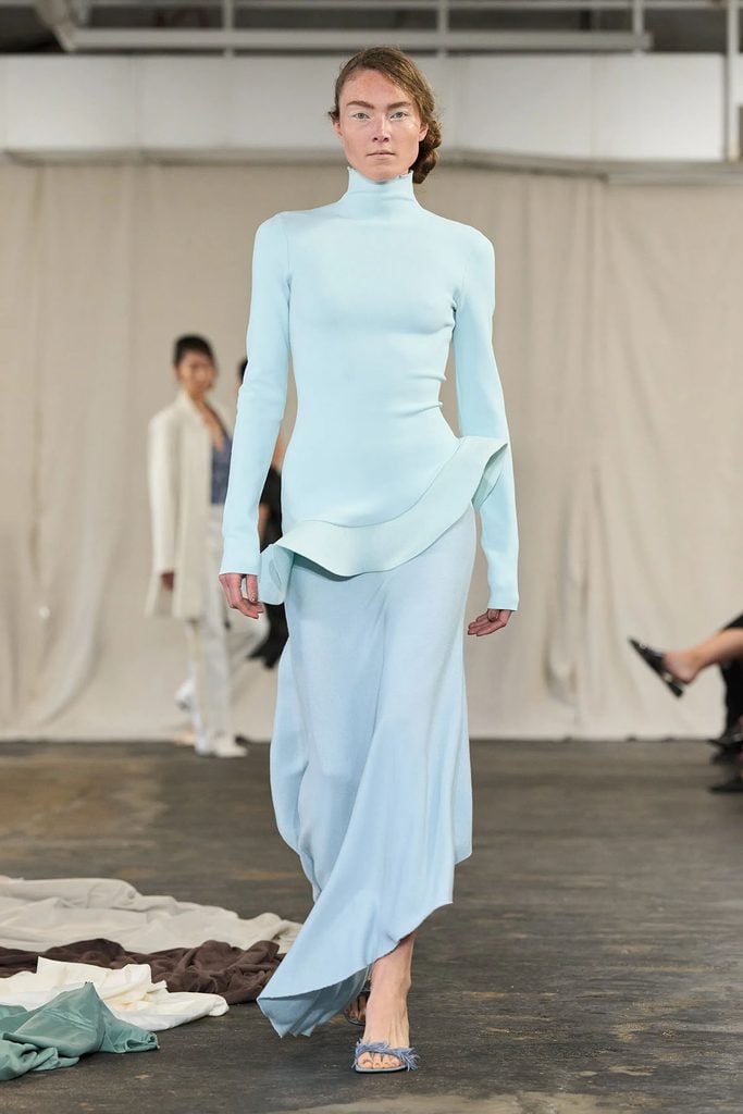 Diotima Spring 2027 runway look featuring a pale blue high-neck top with an asymmetric sculpted hem and a flowing skirt.
