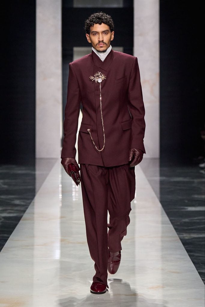 Dolce & Gabbana Fall 2026 menswear runway model wearing a burgundy suit with overlapping lapels, a decorative brooch and a long chain.
