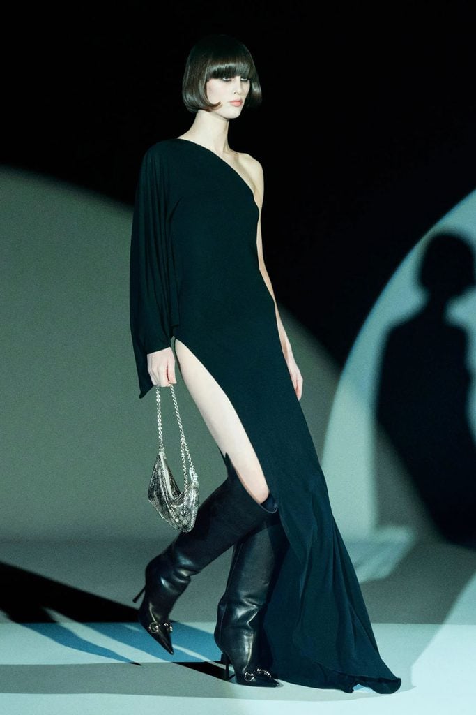 Gucci Pre-Fall 2026 model wearing a black asymmetric jersey gown with a high slit, knee-high leather boots and a silver chain bag.