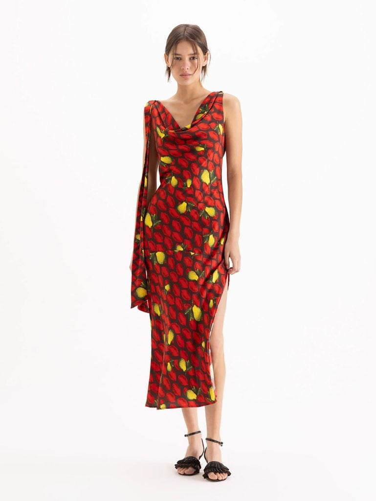Model wearing the Jazmín Chebar Pear dress featuring a red and yellow-green fruit print, draped neckline, long side panel and thigh-high slit.