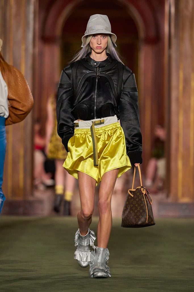 Model wearing a black leather bomber jacket with bright yellow shorts, a grey hat and a monogram bag on the Louis Vuitton Resort 2027 runway.