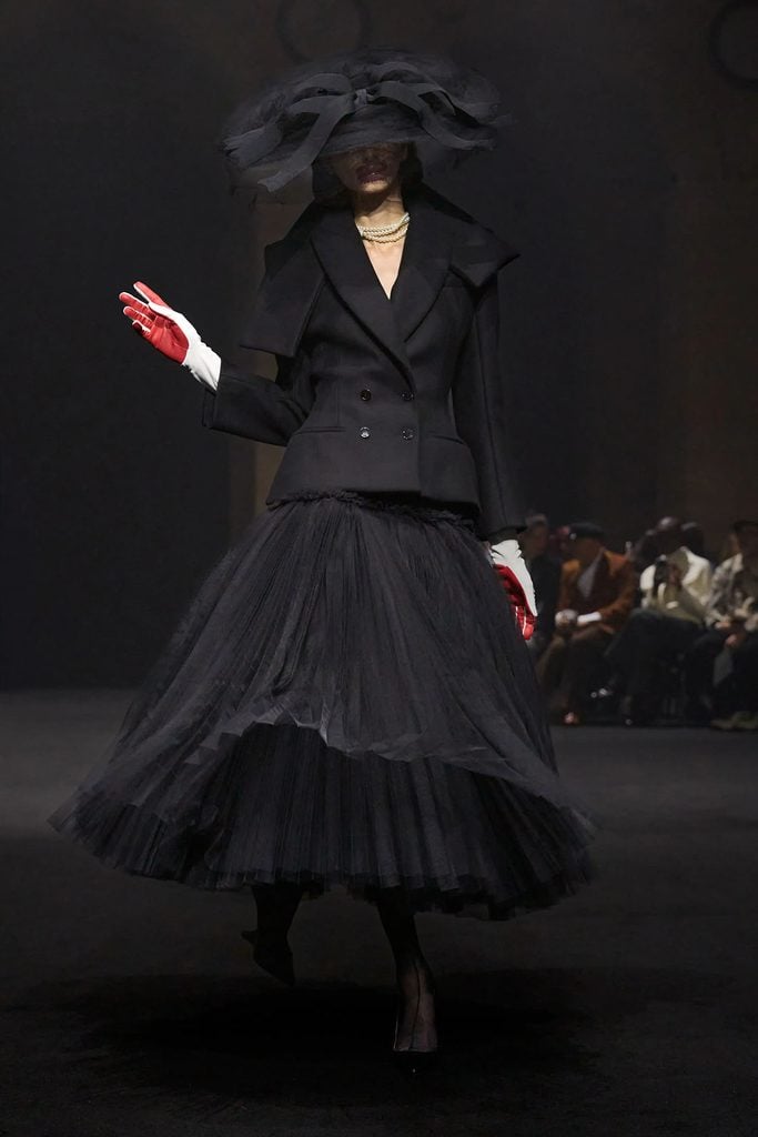 Matières Fécales Fall 2026 runway model wearing a black tailored jacket with a voluminous pleated tulle skirt, oversized hat and red gloves.
