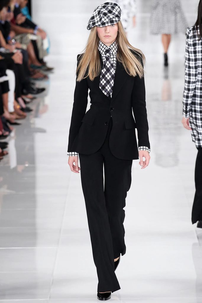 Model wearing a black Ralph Lauren Collection Spring 2014 trouser suit with contrasting checked shirt, tie and matching checked cap on the runway.