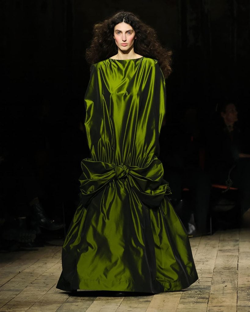 Simone Rocha Fall 2026 runway model wears the Moss Taffeta Bow Cascade Dress in olive green, featuring an oversized silhouette and gathered bow detailing, later worn by Glenn Close to The Hunger Games London premiere.