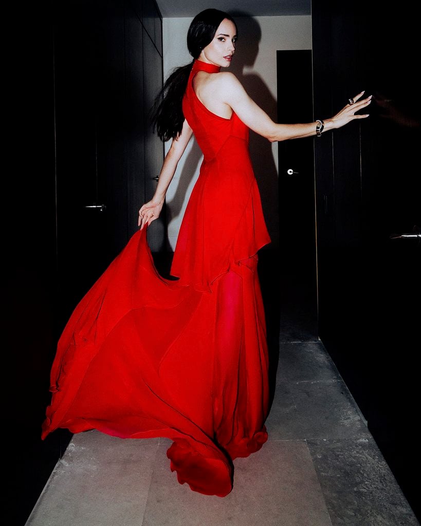 Sofia Carson poses in a hallway wearing her flowing red Lanvin Spring 2027 dress, with its long train spread across the floor.