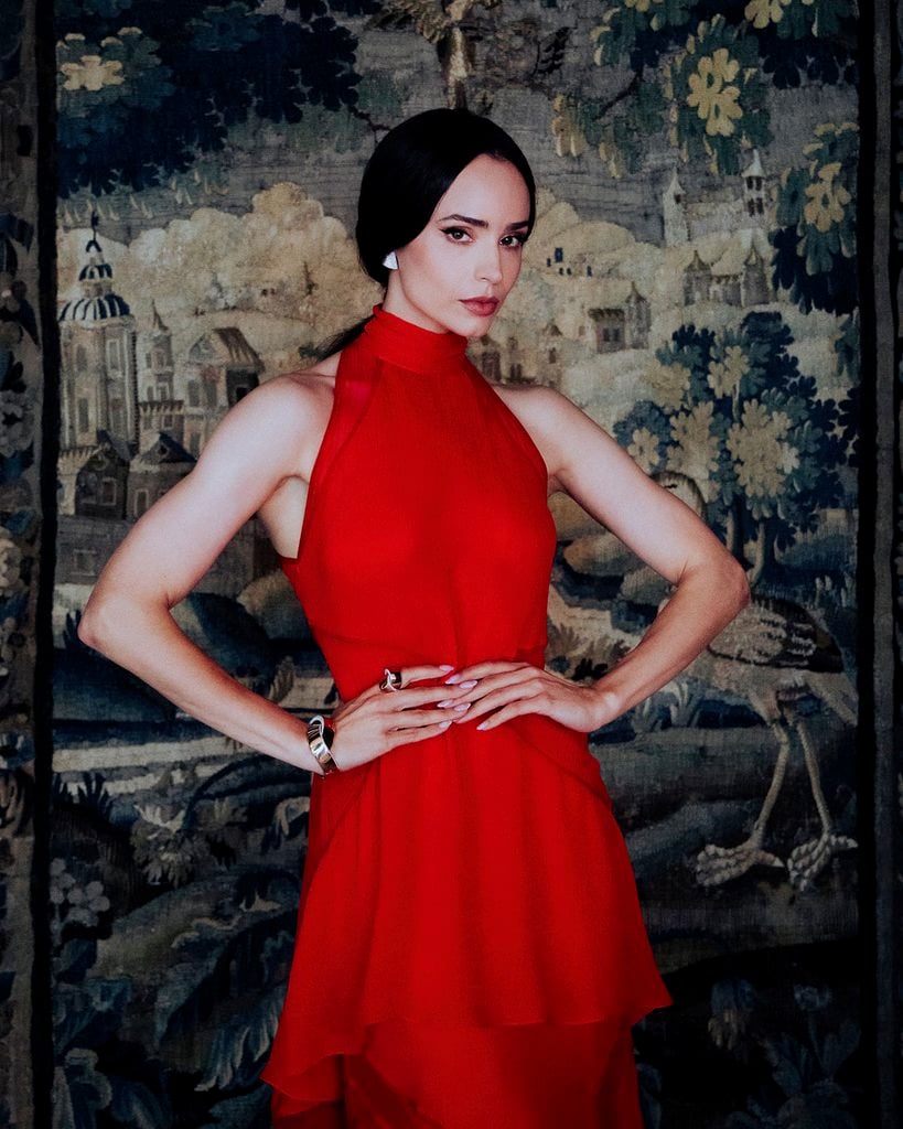 Sofia Carson poses against a decorative tapestry in her red Lanvin halterneck dress, wearing silver earrings, a bracelet and rings.