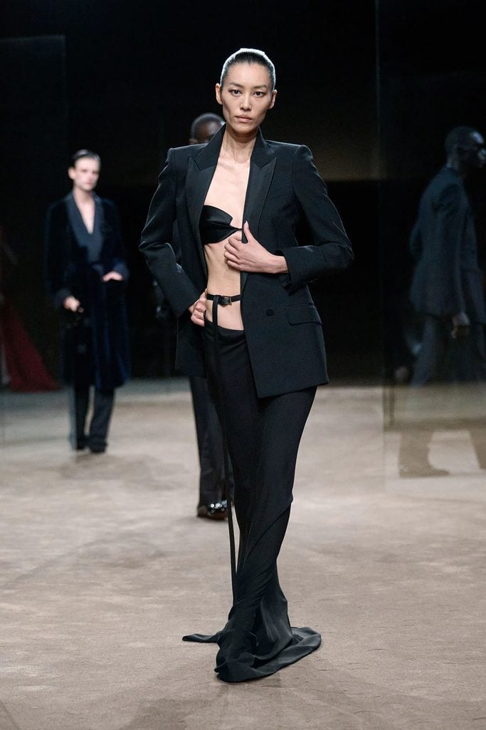Tom Ford Spring 2027 runway model wears a black tuxedo jacket over a bra and draped skirt from Haider Ackermann's collection.