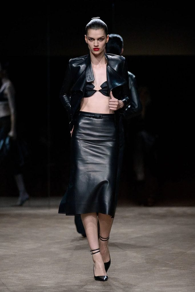 Tom Ford Spring 2027 runway model wearing a cropped black leather officer jacket, sculptural bra top and matching pencil skirt.